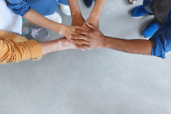 View from above of group of people keeping their hands in pile expressing social friendship, unity, agreement and support. Five people of different sexes collaborating showing their strength
