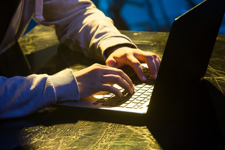 Hooded computer hacker stealing information with laptop on colored studio background