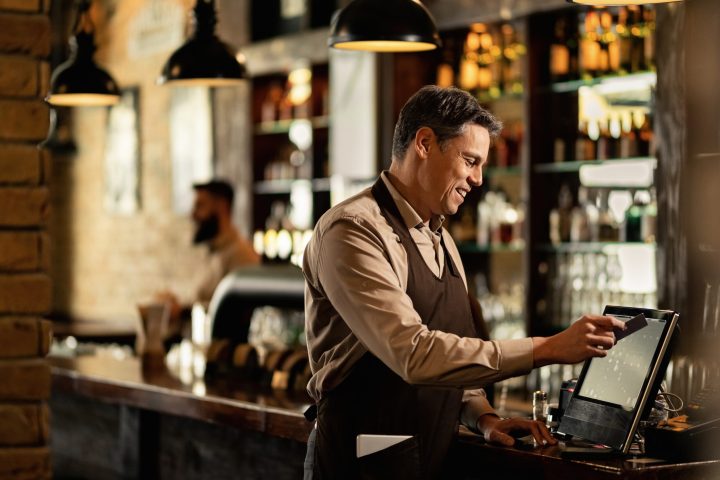 Happy waiter using cash register while checking orders and working in a pub.