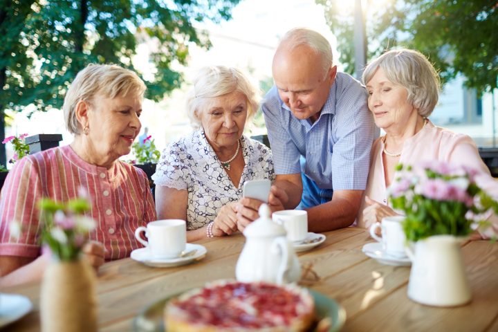 Three senior women and one senior man spending time in outdoors cafe with tea and pastry and watching photos on smartphone