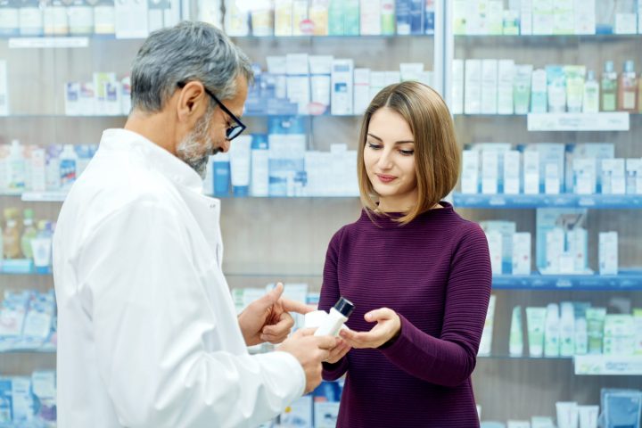 Pharmacist offering pretty female customer medical products in drugstore. Young woman holding white bottles, choosing product. Side view of mature chemist wearing glasses and white lab coat.