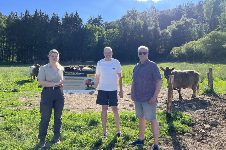 Drei Personen stehen auf einer Wiese mit Kühen, zwei halten ein Schild mit Text zum Altmühltaler Weiderind.